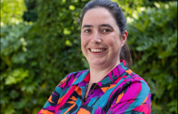 Vrouw met donker haar in een kleurrijk shirt lacht buiten met groene planten op de achtergrond. (vergroot foto)
