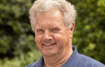Portret van een lachende man met grijs haar, buiten in een groene omgeving. (vergroot foto)