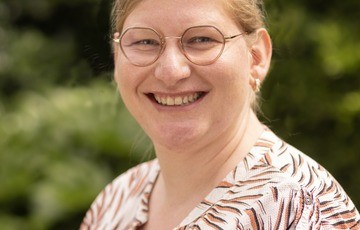 Portret van een lachende vrouw met bril, buiten met groene achtergrond. (vergroot foto)
