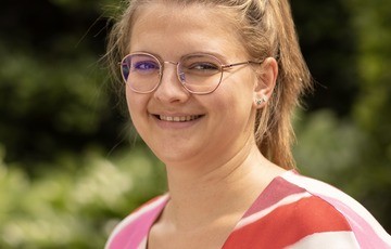 Portret van een lachende vrouw met bril en een rood-wit gestreepte top, buiten in een groene omgeving. (vergroot foto)