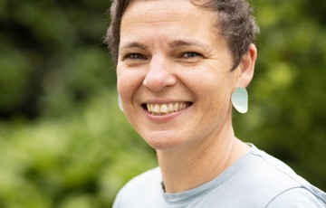 Portret van een vrouw met kort haar en een lichtblauwe top, glimlachend in een groene buitenomgeving. (vergroot foto)
