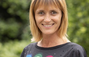 Vrouw met blond haar en een zwarte trui lacht buiten met groene planten op de achtergrond. (vergroot foto)