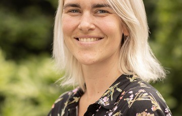 Portret van een vrouw met blond haar en een bloemenblouse, glimlachend in een groene buitenomgeving. (vergroot foto)