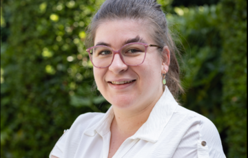 Vrouw met bril en witte blouse lacht, op de achtergrond groene bladeren. (vergroot foto)