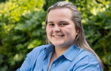 Portret van een lachende vrouw met blond haar, gekleed in een blauw overhemd, voor een groene achtergrond. (vergroot foto)