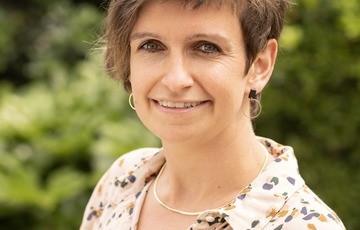 Portret van een vrouw met kort haar, glimlachend, buiten met groene planten op de achtergrond. (vergroot foto)