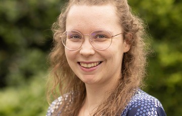 Portret van een lachende vrouw met bril en golvend haar, op een groene, natuurlijke achtergrond. (vergroot foto)