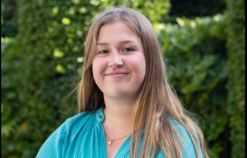 Portret van een jonge vrouw met lang haar, glimlachend, in een groene buitenomgeving. (vergroot foto)